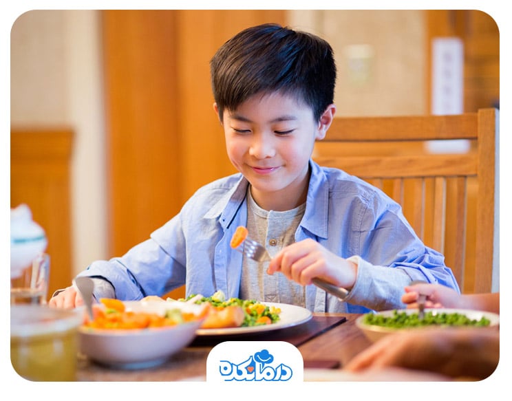 پسری در کنار میز غذا که در حال خوردن هویج و سایر سبزیجات است