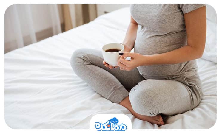 زن بارداری که روی تخت نشسته و در حال خوردن یک فنجان قهوه است