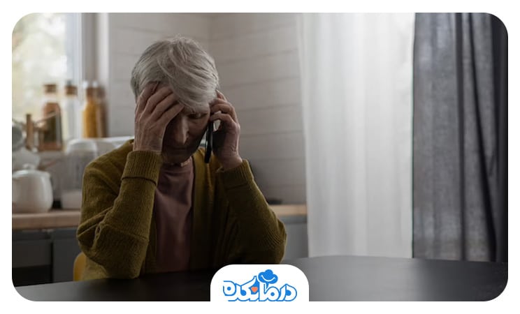 زن سالمند مبتلا به افسردگی اساسی که پشت میز نشسته، سر خود را با دست گرفته و با موبایل صحبت می‌کند