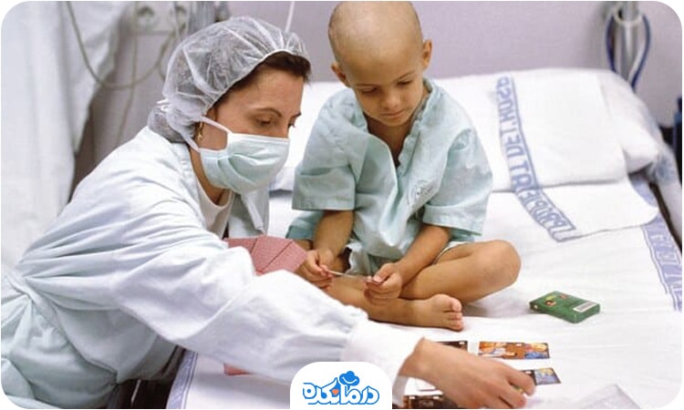 کودک مبتلا به سرطان روی تخت بیمارستان نشسته و انکولوژیست با او بازی می‌کند