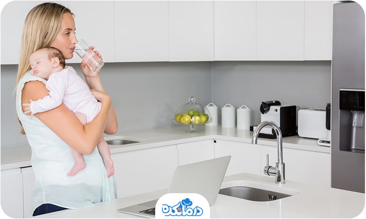 خانمی که نوزادش را در آغوش گرفته است و در حال نوشیدن آب است. مصرف مایعات کافی به درمان یبوست پس از زایمان کمک می‌کند.