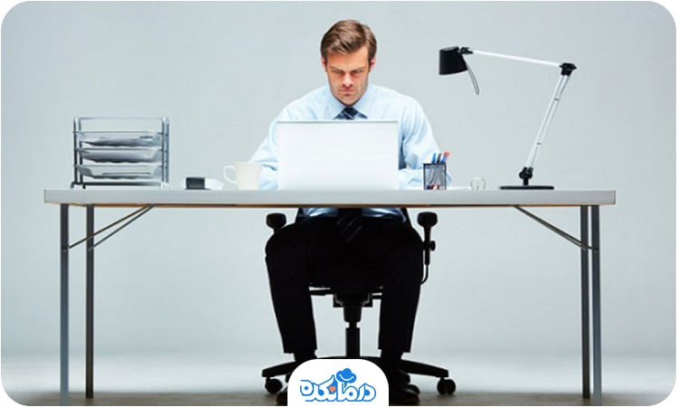 یک مرد پشت میز نشسته و  در حال کار کردن با لپ‌تاپ است