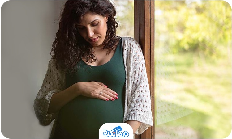 یک زن باردار که کنار پنجره ایستاده و دست خود را روی شکم قرار داده است