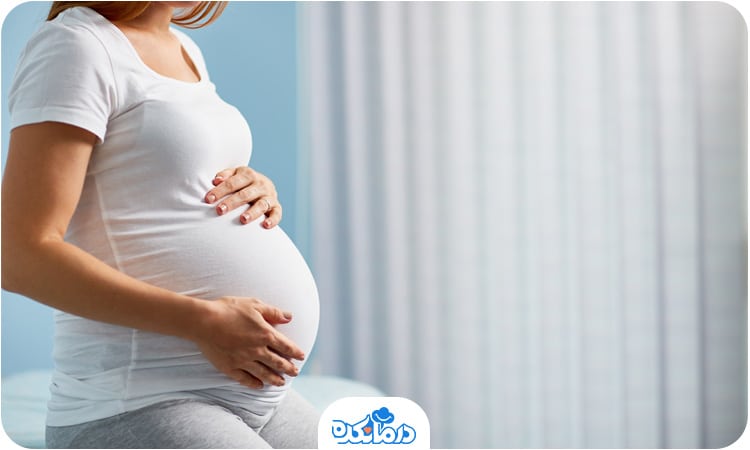 یک زن باردار که ایستاده و ۲ دست خود را روی شکم گذاشته است