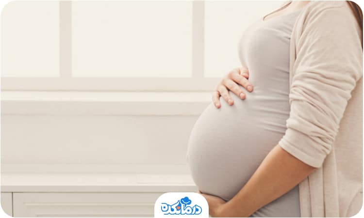 یک زن باردار در کنار پنجره که دست خود را روی شکم قرار داده است