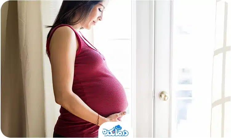  یک زن باردار که کنار پنجره ایستاده است