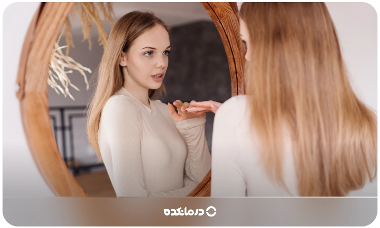 تصویر یک زن جوان که روبروی آینه ایستاده است را می‌بینید
