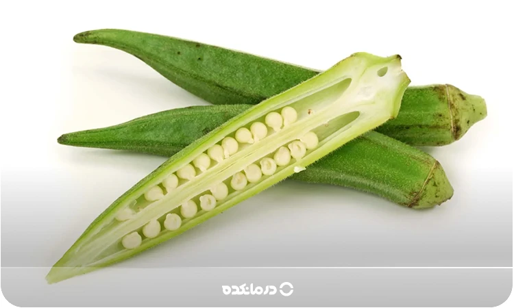 تصویر بامیه سبز درسته و نصف شده که دانهها مشخص هستند را می بینید