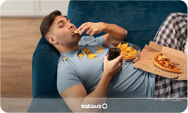 تصویر فردی که روی مبل خوابیده و در حال خوردن غذاست